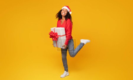 Photo for Joyful black woman in Santa hat, proudly displaying large gift box, posing against vibrant yellow background, symbolizing the festive winter holiday spirit, full length - Royalty Free Image