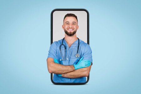 Téléchargez les photos : Jeune médecin européen confiant en uniforme bleu, les bras croisés souriant à la caméra, affiché sur l'écran du smartphone sur fond bleu - en image libre de droit