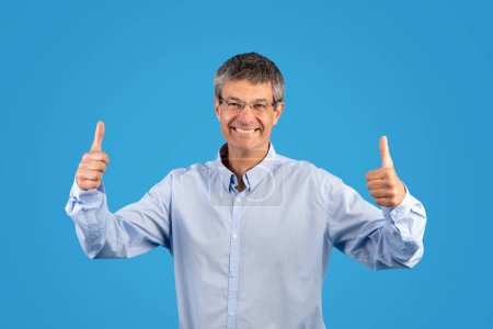 Photo for Happy gray European man in shirt gesturing thumbs up with both hands, smiling posing glasses and approving great business offer with likes standing on blue background. I like it - Royalty Free Image