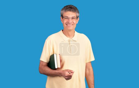 Photo for Knowledgeable mature man with eyeglasses holds books, smiling expressing wisdom and the joy of learning, blue studio backdrop. Education and academic knowledge in any age - Royalty Free Image
