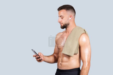 Photo for Calm confident handsome strong millennial european man with beard, naked torso, towel, wireless headphones, chatting on phone, isolated on gray studio background. Body care, sport with music - Royalty Free Image