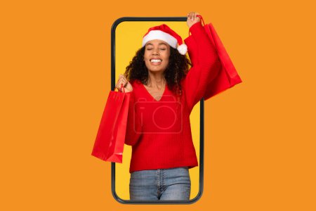 Photo for Merry black lady in Santa hat, showcasing her shopper bags from Christmas sales, framed within the confines of mobile screen on orange background - Royalty Free Image