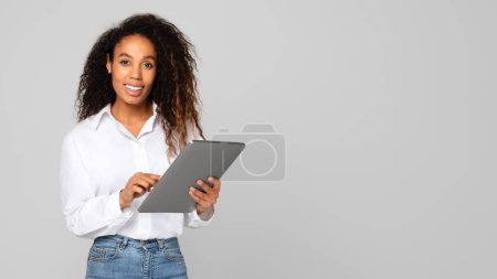 Photo for Black millennial lady using her digital tablet gadget working as freelancer, browsing web while posing near empty space for online offer, standing confidently over gray studio background. Panorama - Royalty Free Image