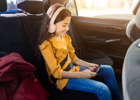 Foto de Alegre chica joven con auriculares disfruta de su teléfono inteligente mientras está sentado en el asiento trasero de los coches, la luz del sol que fluye en, mochila de la escuela roja junto a ella - Imagen libre de derechos