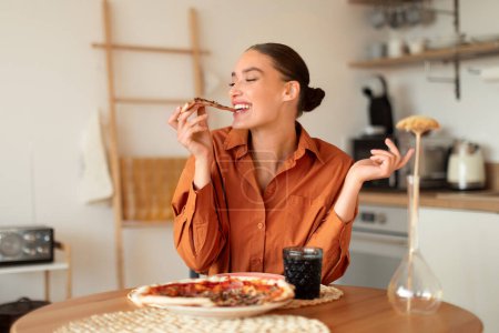 Foto de Joven mujer alegre en blusa naranja disfruta de un bocado de rebanada de pizza, sentado en la cocina moderna con iluminación ambiental y decoración minimalista, espacio libre - Imagen libre de derechos