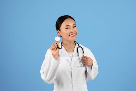 Foto de Retrato de la sonriente mujer médica asiática con estetoscopio en la mano posando sobre fondo azul, trabajador médico profesional que usa uniforme de capa blanca mirando a la cámara, listo para chequear con el paciente - Imagen libre de derechos