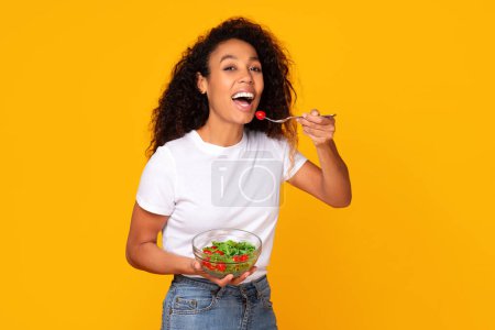 Téléchargez les photos : Jeune femme noire dégustant une salade verte fraîche, souriant sur fond de studio jaune. Lady dans un style de vie décontracté embrassant saine et la nutrition, minceur sur une alimentation saine - en image libre de droit