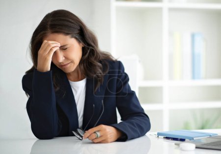 Photo for Exhausted middle aged businesswoman executive manager sitting at workplace at white modern office, rubbing eyes, hold eyeglasses. Overworked lady suffer from migraine, bad vision, have headache - Royalty Free Image