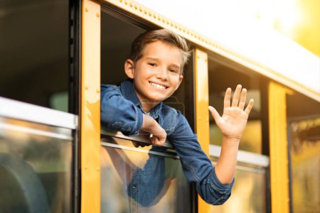 Photo for Happy preteen boy looking out of school bus window and waving hand at camera, cheerful male pupil smiling and saying hello while peeking out of yellow schoolbus, ready for trip, closeup shot - Royalty Free Image