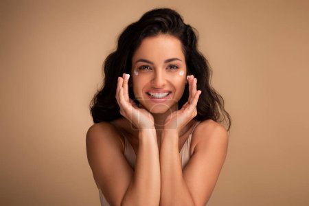 Photo for Happy beautiful indian millennial woman using moisturizing eye cream, attractive brunette lady looking at camera and smiling, enjoying face care routine, beige background - Royalty Free Image