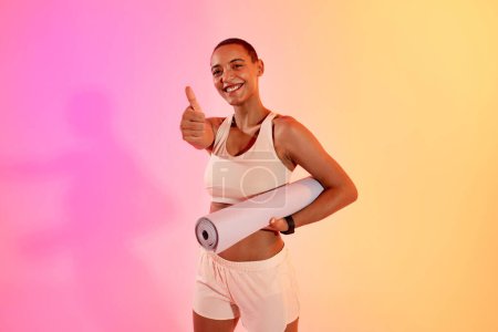 Photo for Smiling pretty young sweat latin woman athlete in sportswear enjoy workout, show thumb up hand sign, hold mat on neon pink studio background. Fitness recommendation, sport and weight loss - Royalty Free Image