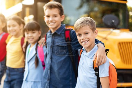 Téléchargez les photos : Heureux groupe multiethnique d'enfants posant à l'extérieur par autobus scolaire jaune, souriants garçons et filles avec des sacs à dos étreignant et regardant la caméra, passer un bon moment ensemble, profiter de leur amitié - en image libre de droit