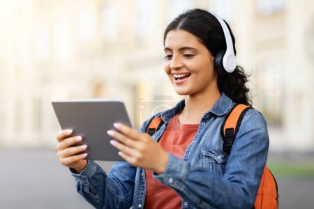 Photo for Happy cheerful pretty young woman student walking by university campus, talking via digital tablet with friend, making plans, using wireless headphones, carrying backpack, copy space - Royalty Free Image
