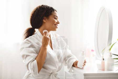 Photo for Beautiful Black Female With Cotton Pads Cleansing Neck Skin At Home, Attractive Young African American Woman Sitting At Dressing Table And Looking In Mirror, Making Daily Beauty Routine - Royalty Free Image