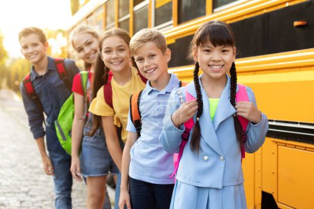 Foto de Retrato de escolares felices posando cerca del autobús escolar amarillo, diversos niños alegres con mochilas sonriendo y mirando a la cámara, listos para ir a casa después del exitoso día de aprendizaje, primer plano - Imagen libre de derechos