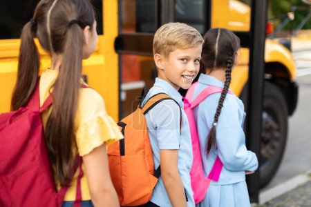 Foto de Primer plano del niño sonriente con la mochila abordando el autobús escolar amarillo, mirando a la cámara, grupo de sus compañeros de clase también subiendo al autobús después de sus lecciones, todo listo para su viaje juntos - Imagen libre de derechos