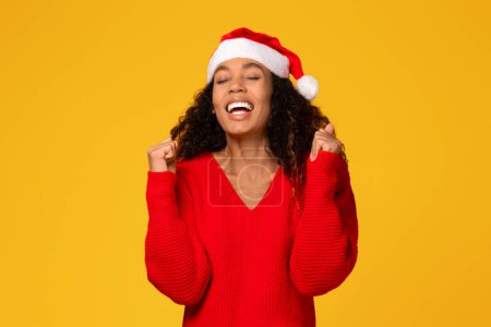 Photo for Joyful young black lady in Santa hat clenching fists and screaming Yes, standing on yellow background, being happy with huge Christmas sales - Royalty Free Image