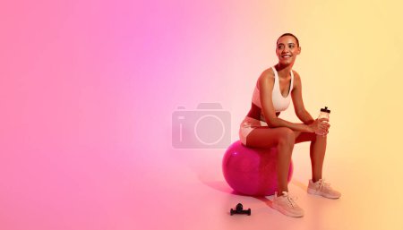 Foto de Mujer latina joven delgada positiva en ropa deportiva sentarse, descansando en la pelota de fitness con botella de agua sobre fondo de estudio de color rosa neón. Estilo de vida, deporte, salud y descanso - Imagen libre de derechos