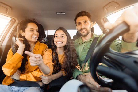 Téléchargez les photos : Famille enthousiaste partage un moment agréable dans leur voiture, avec la fille curieuse soulignant quelque chose d'intéressant, et les parents partageant son enthousiasme avec des expressions joyeuses - en image libre de droit
