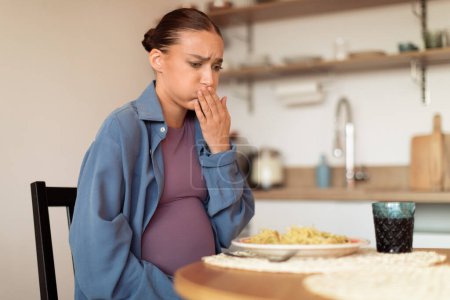 Téléchargez les photos : Femme enceinte s'assoit à sa table de cuisine, montrant des signes de nausées et d'inconfort, peut-être en raison de l'arôme du plat de pâtes en face d'elle, espace libre - en image libre de droit