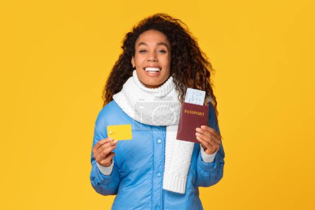 Foto de Joven dama negra sonriente en traje de invierno con tarjeta de crédito, pasaporte y boletos contra fondo amarillo vívido, comprando tour en línea, listo para viajar en invierno - Imagen libre de derechos