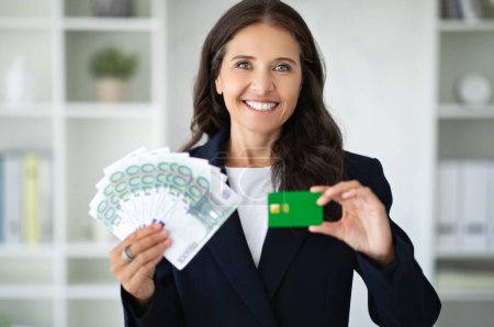 Photo for Positive middle aged european businesswoman recommends credit card, holds a lot of money cash in white office interior. Credit, profit and lifestyle, finance for business, startup, work - Royalty Free Image