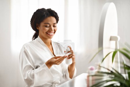Photo for Skincare Routine. Attractive Black Female Applying Makeup Removal Milk On Cotton Pad While Sitting At Dressing Table At Home, Smiling Beautiful Lady Cleansing Skin In The Morning, Copy Space - Royalty Free Image