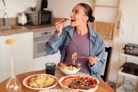 Téléchargez les photos : Joyeuse femme enceinte dans un cadre de cuisine confortable, prenant avec impatience une bouchée de pizza tout en ayant des pâtes et une salade à proximité, appréciant diverses saveurs pendant sa grossesse - en image libre de droit