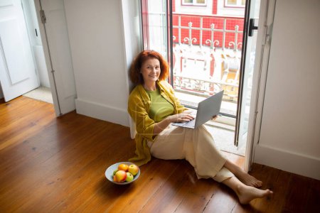 Téléchargez les photos : Femme âgée souriante utilisant un ordinateur portable tout en étant assise sur le sol par la fenêtre, femme âgée heureuse engagée dans le travail ou les loisirs dans sa maison ou sa chambre d'hôtel confortable pendant ses années de retraite, copier l'espace - en image libre de droit