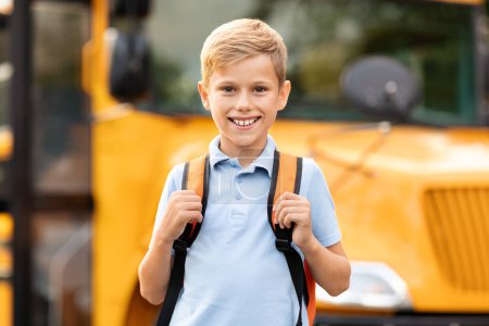 Foto de Sonriendo lindo niño preadolescente posando cerca de autobús escolar amarillo al aire libre, niño varón feliz con mochila de pie fuera y mirando a la cámara, listo para las clases, colegial disfrutando de estudio, espacio de copia - Imagen libre de derechos