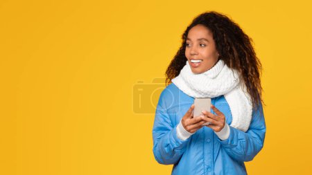 Téléchargez les photos : Joyeuse femme vêtue d'une veste d'hiver bleue et d'une écharpe blanche, tenant un smartphone avec les deux mains, debout sur un fond jaune vibrant, regardant de côté l'espace libre et souriant - en image libre de droit