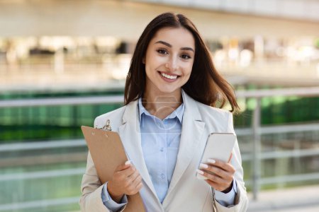 Photo for Smiling pretty confident young european lady in suit with clipboard, enjoy work, typing on smartphone in city outdoors. Business, startup, device and lifestyle, app for chat - Royalty Free Image