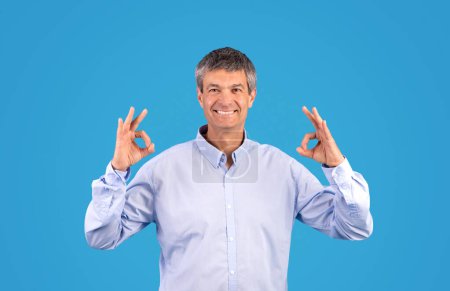 Téléchargez les photos : Heureux homme européen mature en chemise montrant OK signe avec les deux mains souriant en regardant la caméra, approuvant belle offre d'affaires debout en studio sur fond bleu. Tout va bien concept - en image libre de droit