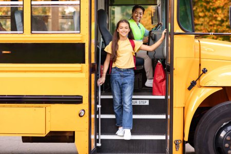 Foto de Feliz niña preadolescente bajándose del autobús escolar amarillo, alegre niña bajando del vehículo, amigable conductora negra de uniforme mirándola y sonriendo, espacio libre - Imagen libre de derechos