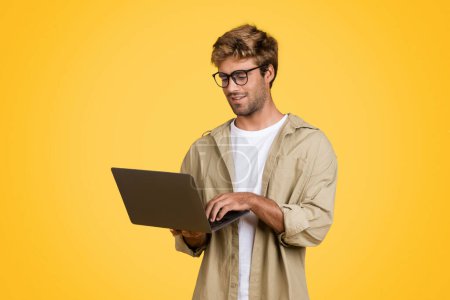 Photo for Stylish young European man is engrossed in using laptop, diligently typing on keyboard, stands poised against distinct yellow studio backdrop, focused on his task - Royalty Free Image