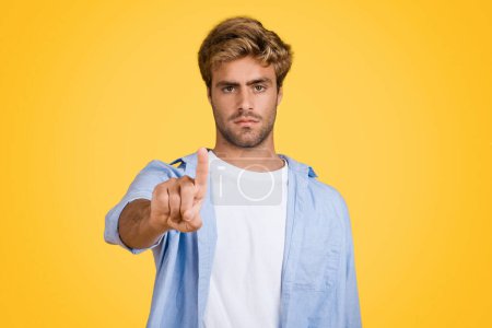 Photo for Focused young European man interacts with unseen digital screen or button, standing against vibrant yellow studio backdrop. A nod to cutting-edge modern technologies - Royalty Free Image