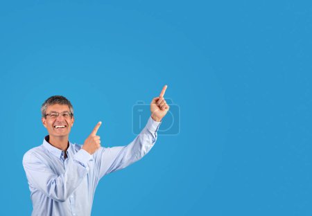 Photo for Excited businessman in shirt pointing to blank space aside with joyful expression, standing against blue studio backdrop, beaming with contagious enthusiasm. Banner for promotional content - Royalty Free Image