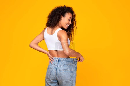 Téléchargez les photos : Femme afro-américaine démontre sa taille parfaite après une grande perte de poids, portant de gros jeans, se tenant à l'arrière de la caméra sur fond de studio jaune. Concept de motivation minceur - en image libre de droit