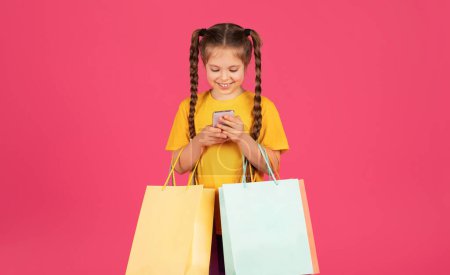 Photo for Shopping App. Cheerful Little Girl Holding Smartphone And Colorful Paper Shopper Bags, Happy Preteen Female Child Using Mobile Application For Discounts And Offers, Standing On Pink Background - Royalty Free Image