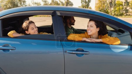 Téléchargez les photos : Mère et fille joyeuses jettent un coup d'oeil hors de leur voiture, profitant de la vue, tandis que le père conduit, tous partageant moment lumineux ensemble pendant la journée ensoleillée - en image libre de droit
