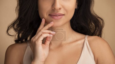 Téléchargez les photos : Recadrée de belle jeune femme touchant le visage, démontrant une peau parfaite, des lèvres douces, un maquillage nu isolé sur fond studio beige. Soins de la peau soins de santé procédures cosmétiques concept - en image libre de droit