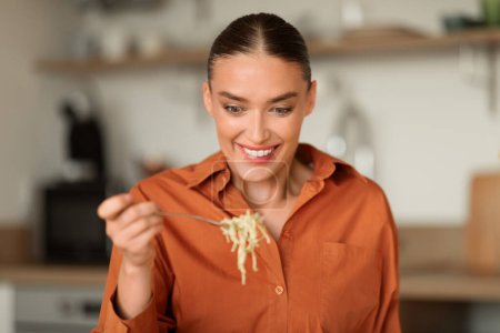 Photo for Happy woman sits in contemporary kitchen, gleefully savoring forkful of noodles, with ambient kitchen details subtly present in the background - Royalty Free Image