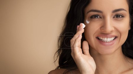 Photo for Anti-aging eye care cosmetics concept. Happy attractive brunette long-haired millennial indian woman applying eye cream for smooth skin, closeup, beige studio background, panorama with copy space - Royalty Free Image