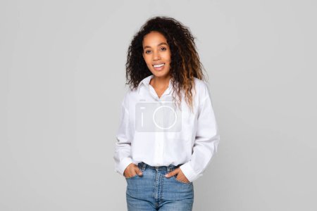 Téléchargez les photos : Jeune femme afro-américaine en jean denim et chemise blanche, souriant avec confiance en se tenant la main dans des poches sur fond de studio gris. Mode moderne et concept de beauté - en image libre de droit