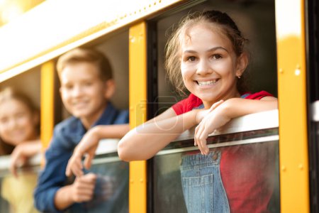 Téléchargez les photos : Groupe d'enfants regardant par la fenêtre de l'autobus scolaire, souriant à la caméra, camarades de classe joyeux profiter de leur voyage en autobus scolaire jaune, heureux de conduire un véhicule ensemble, accent sélectif sur la jeune fille - en image libre de droit