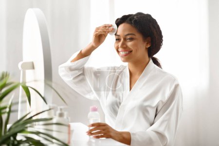 Photo for Beautiful Black Woman Cleansing Skin With Micellar Water And Cotton Pad While Making Daily Beauty Routine At Home, Attractive African American Female Enjoying Skincare Treatments, Copy Space - Royalty Free Image