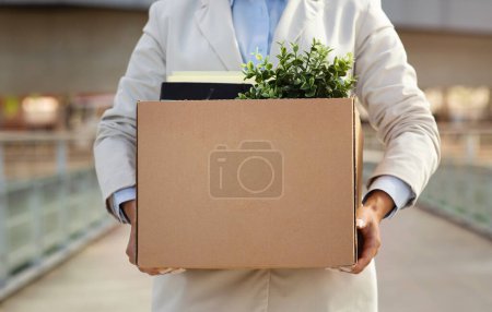 Photo for Millennial caucasian lady manager professional hold cardboard box filled with office essentials, including green potted plant, in city outdoors. Problems with work, dismissal, quit - Royalty Free Image