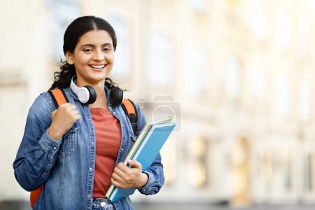 Téléchargez les photos : Bourses d'études collégiales pour étudiants étrangers. Jeune femme indienne heureuse avec sac à dos et cahiers posant en plein air sur le campus, se reposant après les cours, regardant et souriant à la caméra, Copier l'espace - en image libre de droit