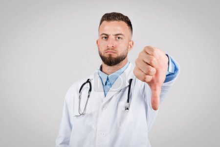 Photo for European male doctor in white coat giving thumbs-down sign, expressing disapproval or negative feedback, standing against grey background - Royalty Free Image