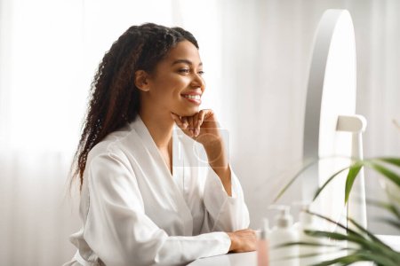 Photo for Beauty Secrets. Attractive Black Woman Looking In Mirror While Sitting At Dressing Table At Home, Happy Beautiful African American Female Touching Chin, Smiling To Her Reflection, Copy Space - Royalty Free Image